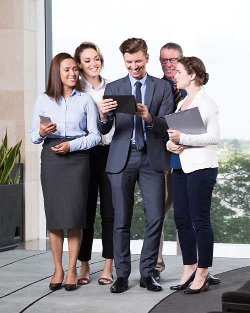 Group of five cheerful business people standing in office lobby, looking at digital tablet computer in his young leaders hands and smiling